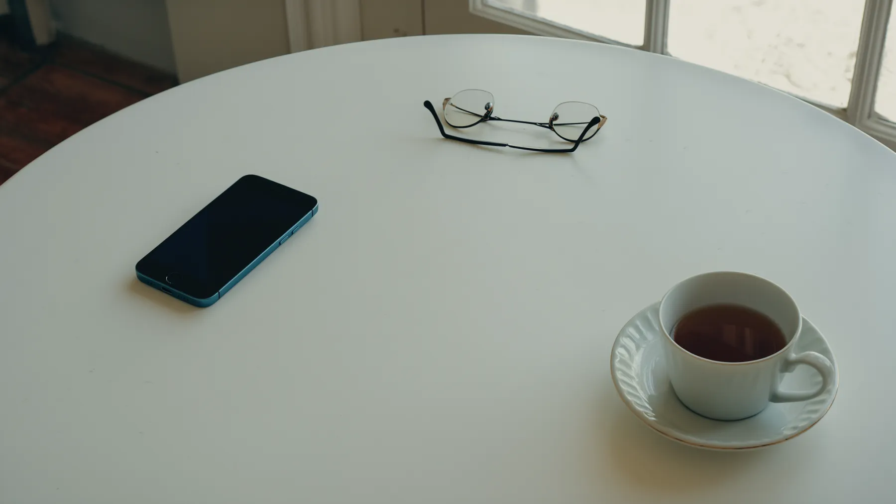 A phone on a kitchen table beside a cup of tea and reading glasses
